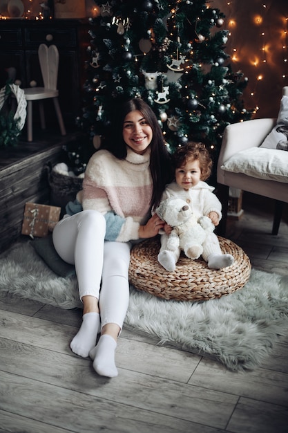 Stock photo of smiling attractive mother and baby sitting under decorated Christmas tree at home.