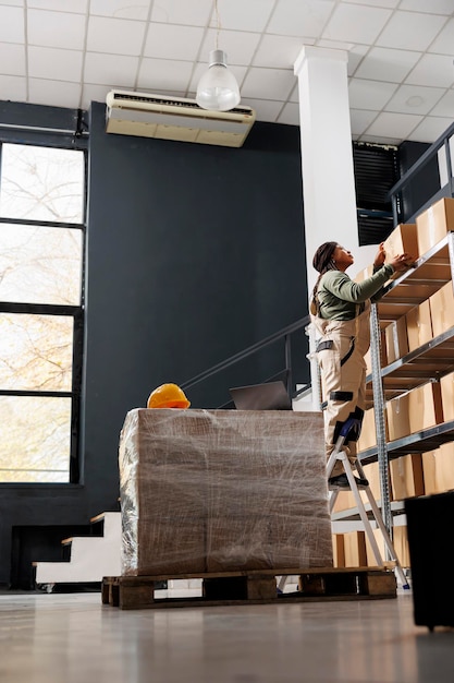 Free photo stockroom worker standing on ladder holding cardboard box, working at shipping products in storehouse. storage room employee preparing customers orders, checking packages in warehouse