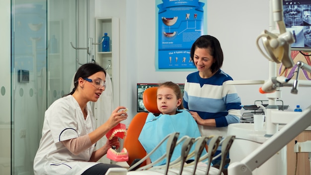 Stomatologist explaining proper dental hygiene using presentation teeth skeleton, extracting a mass from it. Dentist telling to kid the procedure holding sample of human jaw in stomatology office.