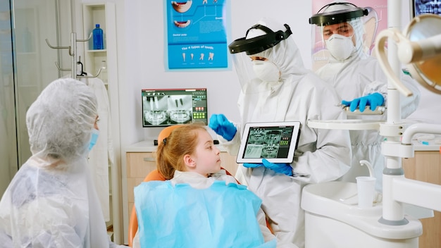 Stomatologist in protective equipment showing on tablet dental x-ray reviewing it with mother of patient. Medical team wearing face shield mask, gloves, explaining radiography using notebook display