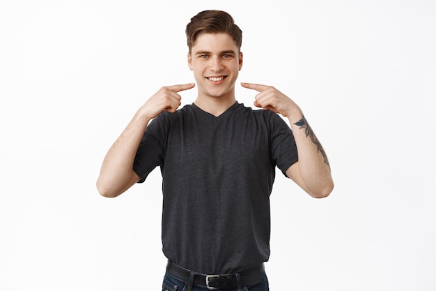 Free photo stomatology and perfect smile concept. handsome young man showing his white teeth, pointing fingers at mouth with satisfied smiling face expression, white background