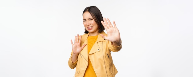 Stop get away from me back off concept Young disgusted asian woman being blinded by smth stretching out hands in defense blocking light standing over white background