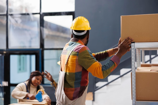 Storehouse manager standing on ladder while reaching for cardboard box. African american warehouse loader wearing safery hard hat and overall workwear unloading shelf with heavy parcels