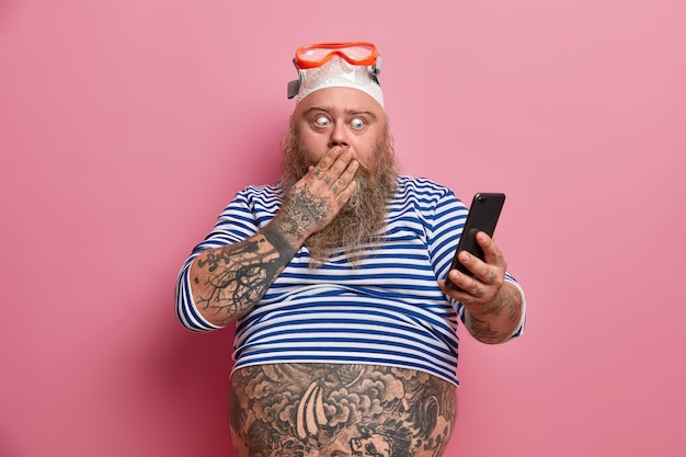 Free Photo stout shocked bearded man covers mouth and stares at smartphone, reads something astonishing, dressed in sailor shirt, swimming goggles, has tattooed belly