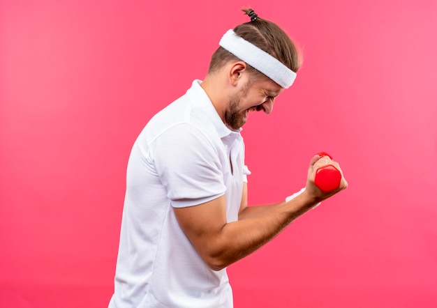 Strained young handsome sporty man wearing headband and wristbands standing in profile view holding dumbbells with closed eyes isolated on pink space