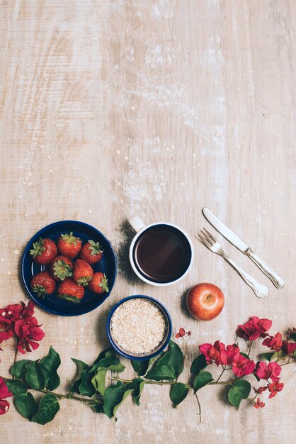 Strawberries; coffee cup; oats; whole apple and cutlery with bougainvillea flowers on wooden backdrop