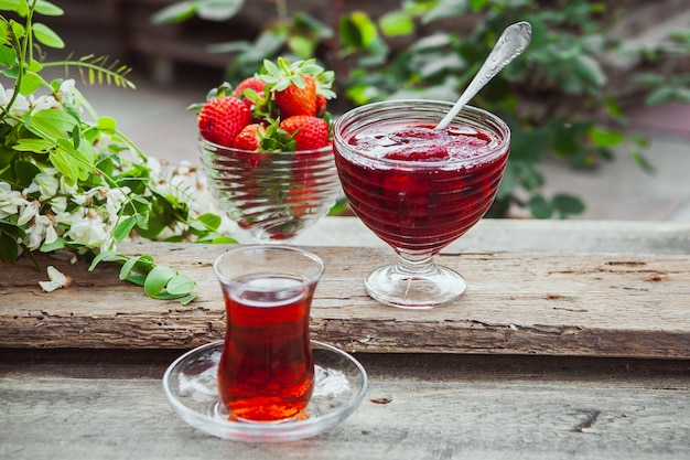 Strawberry jam in a plate with spoon, a glass of tea, strawberries, plant top view on wooden and pavement table