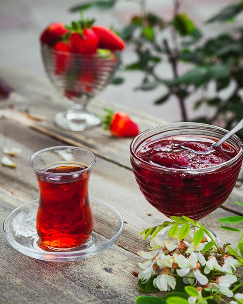 Strawberry jam with a glass of tea, spoon, strawberries, plants in a plate on wooden and pavement table, high angle view.