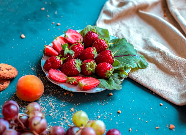 Strawberry plate with mixed fruits around on a blue table .