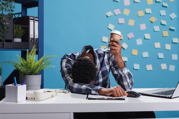 Stressed and fatigued team leader with burnout syndrome sitting in modern office workspace. Tired and exhausted agency office employee falling asleep at desk because of overtime work