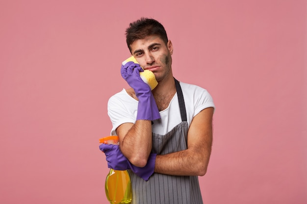 Stressed husband doing housework washing looking tired and desperate. Dirty man wearing apron and gloves holding sponge on cheek and detergent in hand looking tired