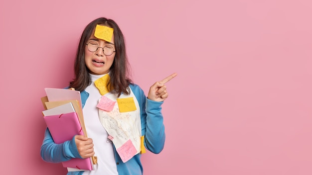 Free photo stressed unhappy female student cries from desprair being fed up of exam preparation points away on blank space overloaded with papers has to remember much information.