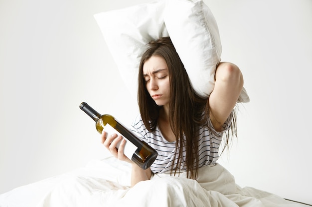 Stressed unhappy woman with messy loose hair, suffering from headache after party, holding pillow and empty bottle