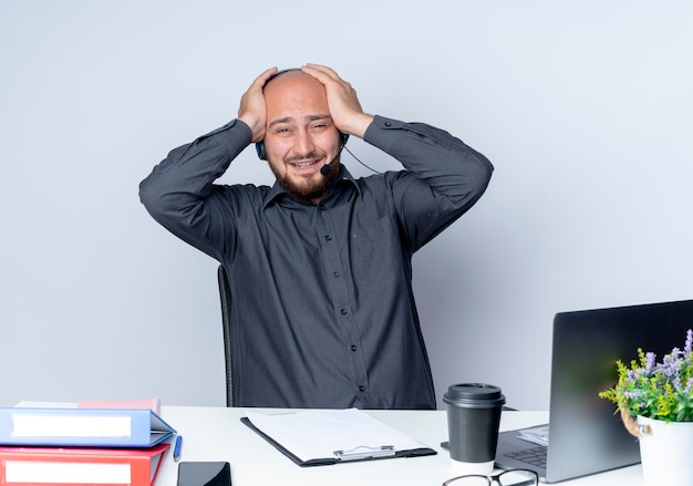 Stressed young bald call center man wearing headset sitting at desk with work tools holding his head isolated on white wall