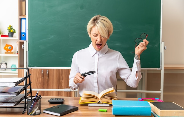 Free photo stressed young blonde female teacher sitting at desk with school tools in classroom holding magnifying glass above open book taking off glasses screaming with closed eyes