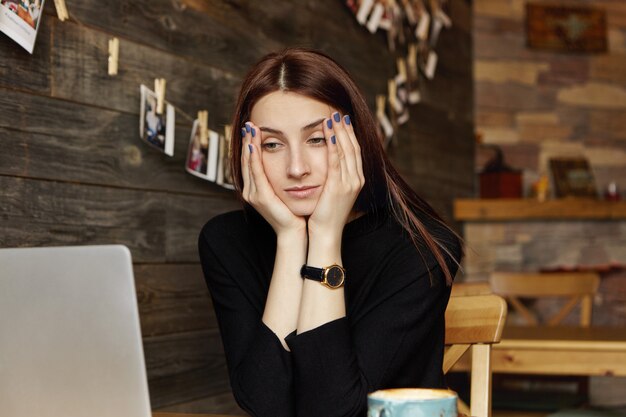 Stressed young Caucasian female freelancer resting face on her hands looking at laptop screen in front of her with bored expression, feeling tired while working remotely at cafe. People and lifestyle