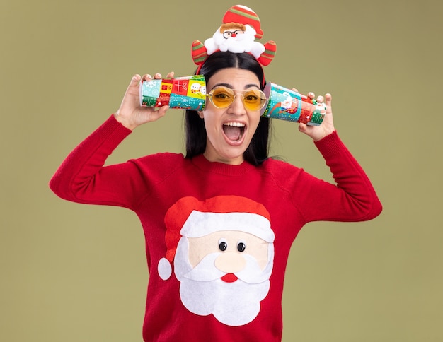 Free Photo stressed young caucasian girl wearing santa claus headband and sweater with glasses holding plastic christmas cups next to ears listening to conversations  screaming isolated on olive green wall