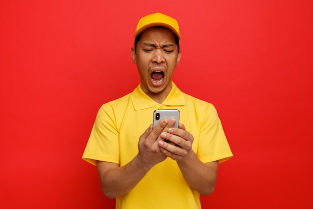 Stressed young delivery man wearing cap and uniform holding and looking at mobile phone screaming 