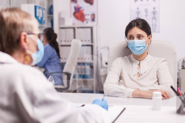 Stressed young patient with face mask against covid-19 during consultation with senior doctor in hospital office. Terrified woman.