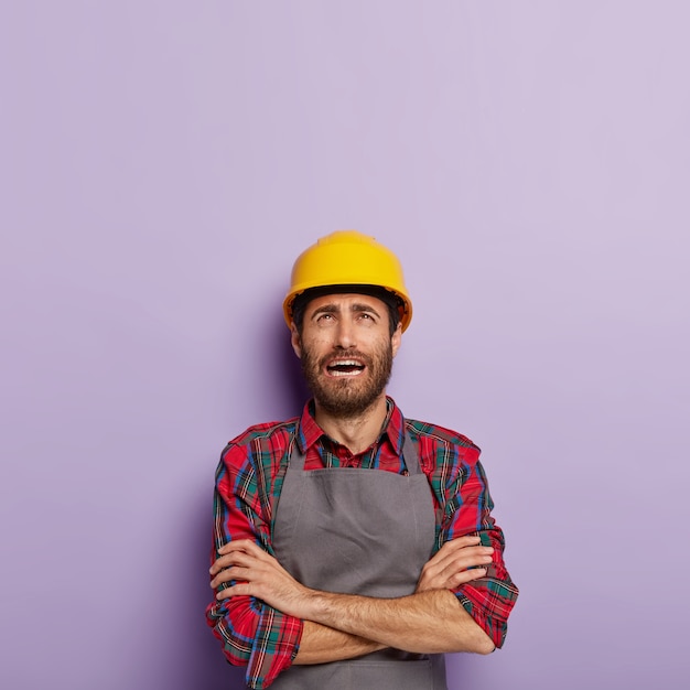 Free Photo stressful dissatisfied male engineer keeps arms folded has miserable face expression, wears checkered shirt and apron, focused above, gets much duties from boss tired of manual work isolated on purple