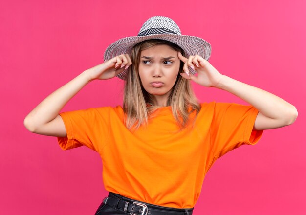 A stressful pretty young woman in an orange t-shirt wearing sunhat pointing at her head while looking side on a pink wall