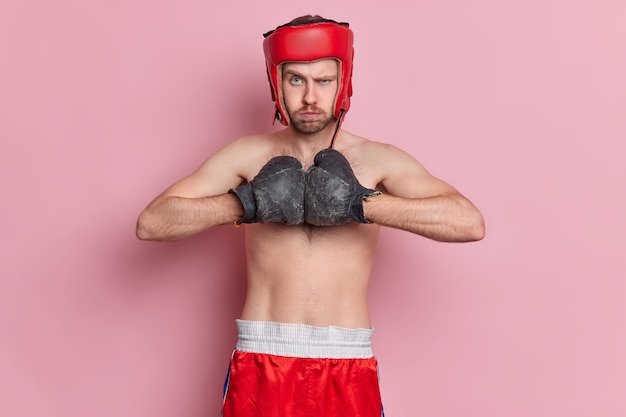 Free photo strict angry unshaven male boxer holds hands together in boxing gloves ready for struggle wears protective helmet and shorts has bare torso