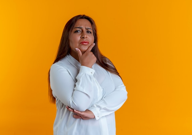 Strict casual caucasian middle-aged woman putting hand under chin isolated on yellow wall with copy space