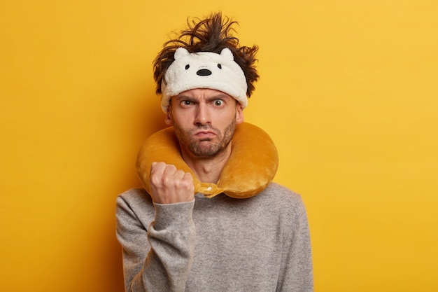 Free photo strict displeased man traveler shows fist, warns you about revenge, wears inflated travel pillow, angry because of airplane delay