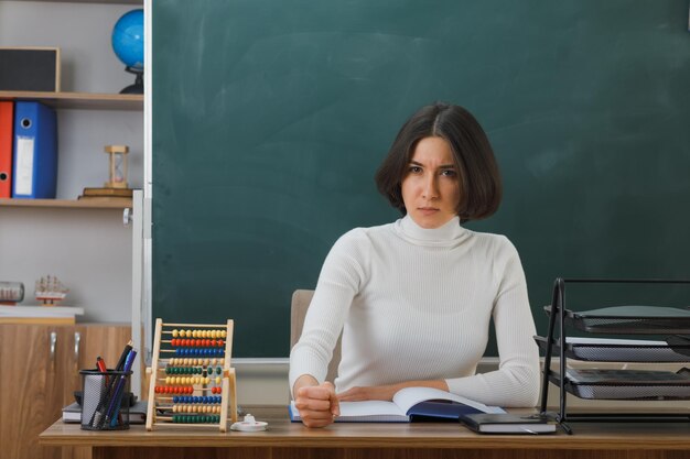 strict looking at camera young female teacher sitting at desk with school tools on in classroom