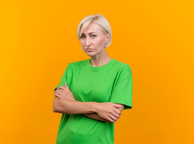 Strict middle-aged blonde slavic woman standing with closed posture looking at camera isolated on yellow background with copy space