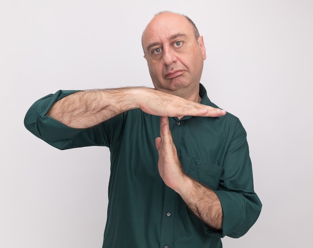 Strict middle-aged man wearing green t-shirt showing timeout gesture isolated on white wall