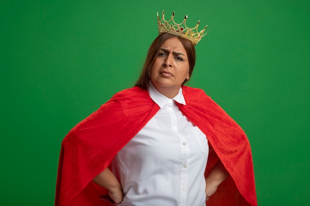 Strict middle-aged superhero female wearing crown putting hands on hip isolated on green background