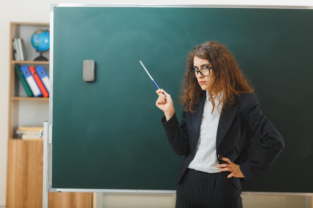 strict putting hand on hips young female teacher standing in front blackboard points at blackboard with pointer in classroom