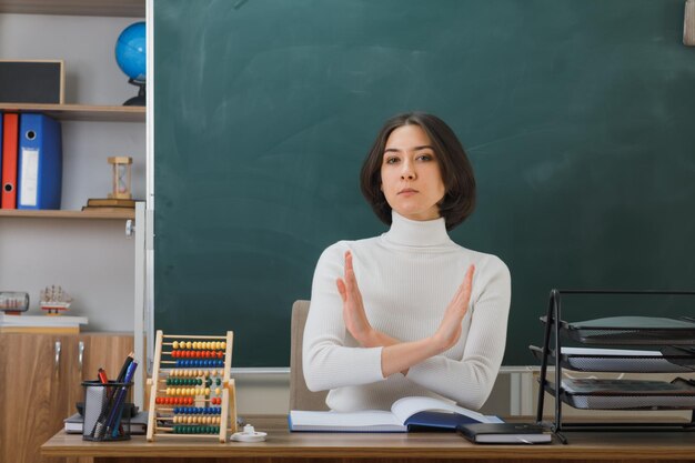 strict showing no gesture young female teacher sitting at desk with school tools on in classroom