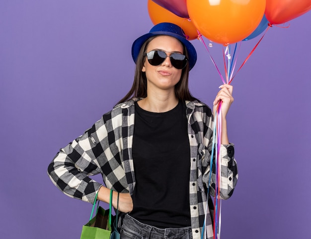 Strict young beautiful girl wearing party hat with glasses holding balloons putting hand on hip 