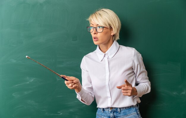 Strict young blonde female teacher wearing glasses in classroom standing in front of chalkboard looking and pointing at side with pointer stick and finger