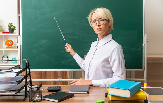 strict young blonde female teacher wearing glasses sitting at desk with school supplies in classroom pointing at chalkboard with pointer stick looking at front