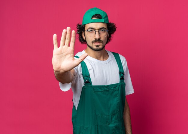 Strict young builder man wearing uniform with cap showing stop gesture 