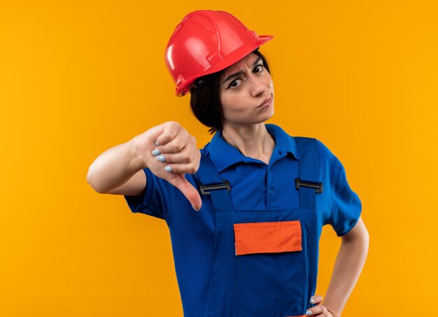 Strict  young builder woman in uniform showing thumb down putting hand on hip isolated on yellow wall