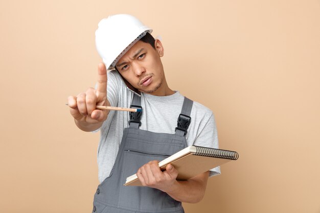 Strict young construction worker wearing safety helmet and uniform holding notepad and pencil doing hold on gesture 