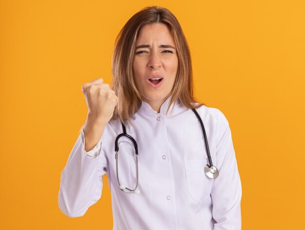 Strict young female doctor wearing medical robe with stethoscope holding fist isolated on yellow wall