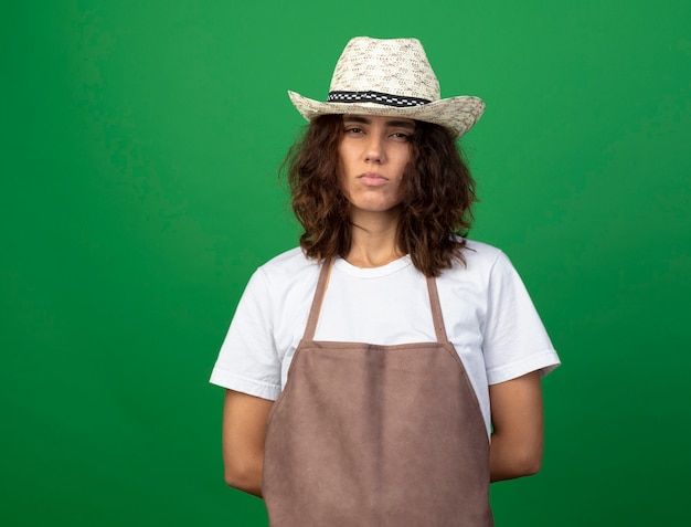 Strict young female gardener in uniform wearing gardening hat keeping hands on waist