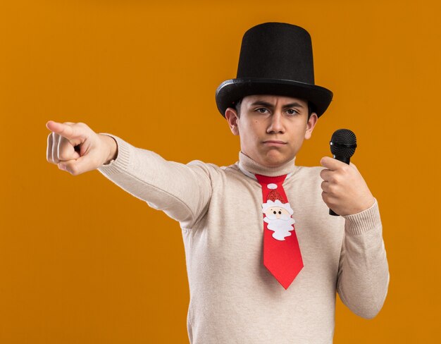 Strict young guy wearing hat with christmas tie holding microphone points at side isolated on yellow wall