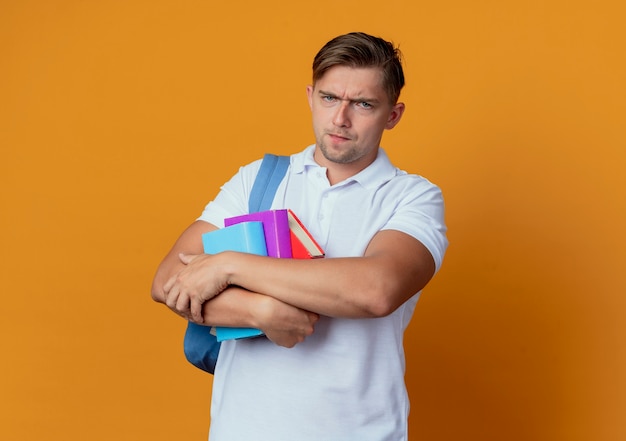 Strict young handsome male student wearing back bag holding books isolated on orange