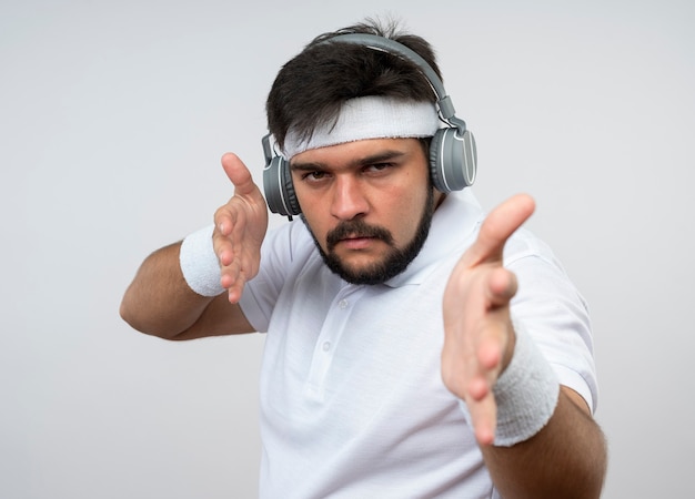 Strict young sporty man wearing headband and wristband with headphones holding out hands  isolated on white wall