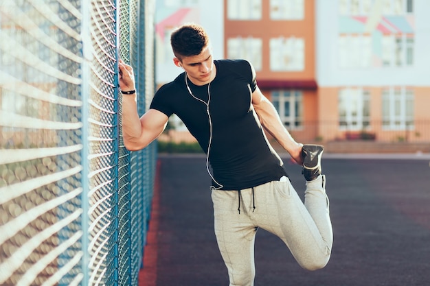 Free photo strong guy with a muscular body at workout near fence in the morning on stadium. he wears sport clothes, listening to music with headphones.