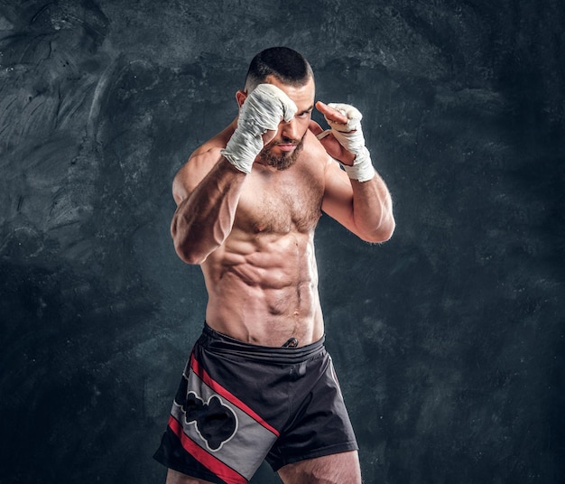 Strong muscular fighter is showing his punch while posing for photographer at dark photo studio.