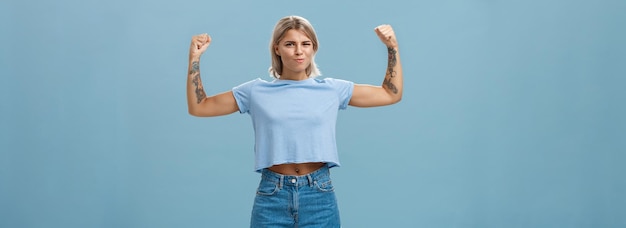 Free Photo strong and powerful goodlooking sportswomen with tattoos in tshirt and shorts raising arms showing big muscles and biceps smiling proudly while bragging with physical strength over blue wall