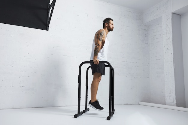 Free photo strong tattooed in white unlabeled tank t-shirt male athlete shows calisthenic moves holding position on parallel bars before classic dips, looking straight