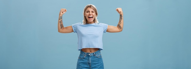 Strong women stand up and fight for rights Portrait of happy entertained and cool young female athletic blonde with tattoos winking and smiling showing muscles and biceps over blue wall